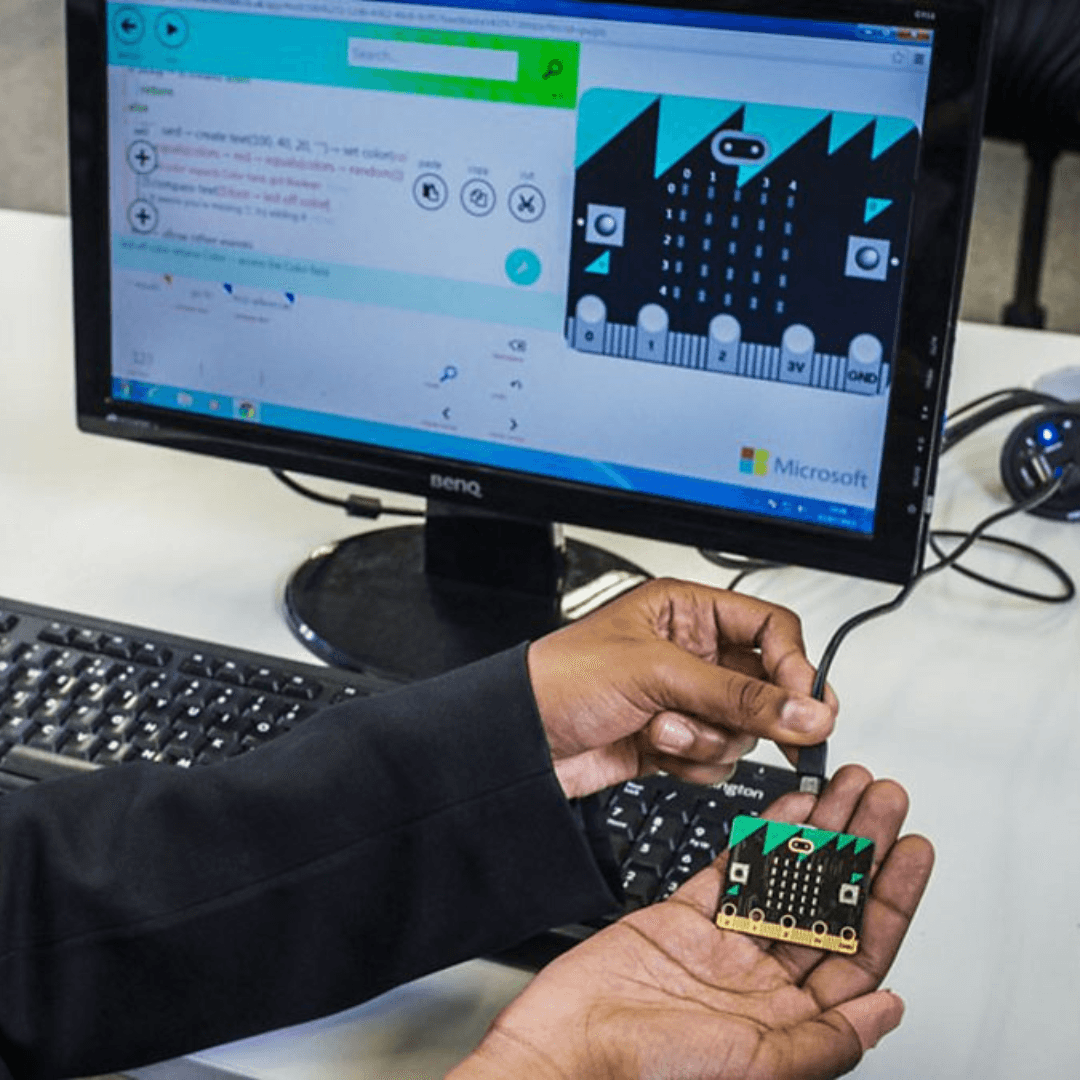 Hand holding a micro:bit in front of a computer showing the micro:bit interface