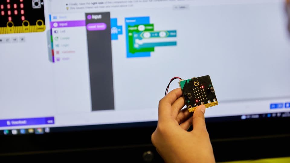 Hand holding a micro:bit in front of a MakeCode block-coding screen