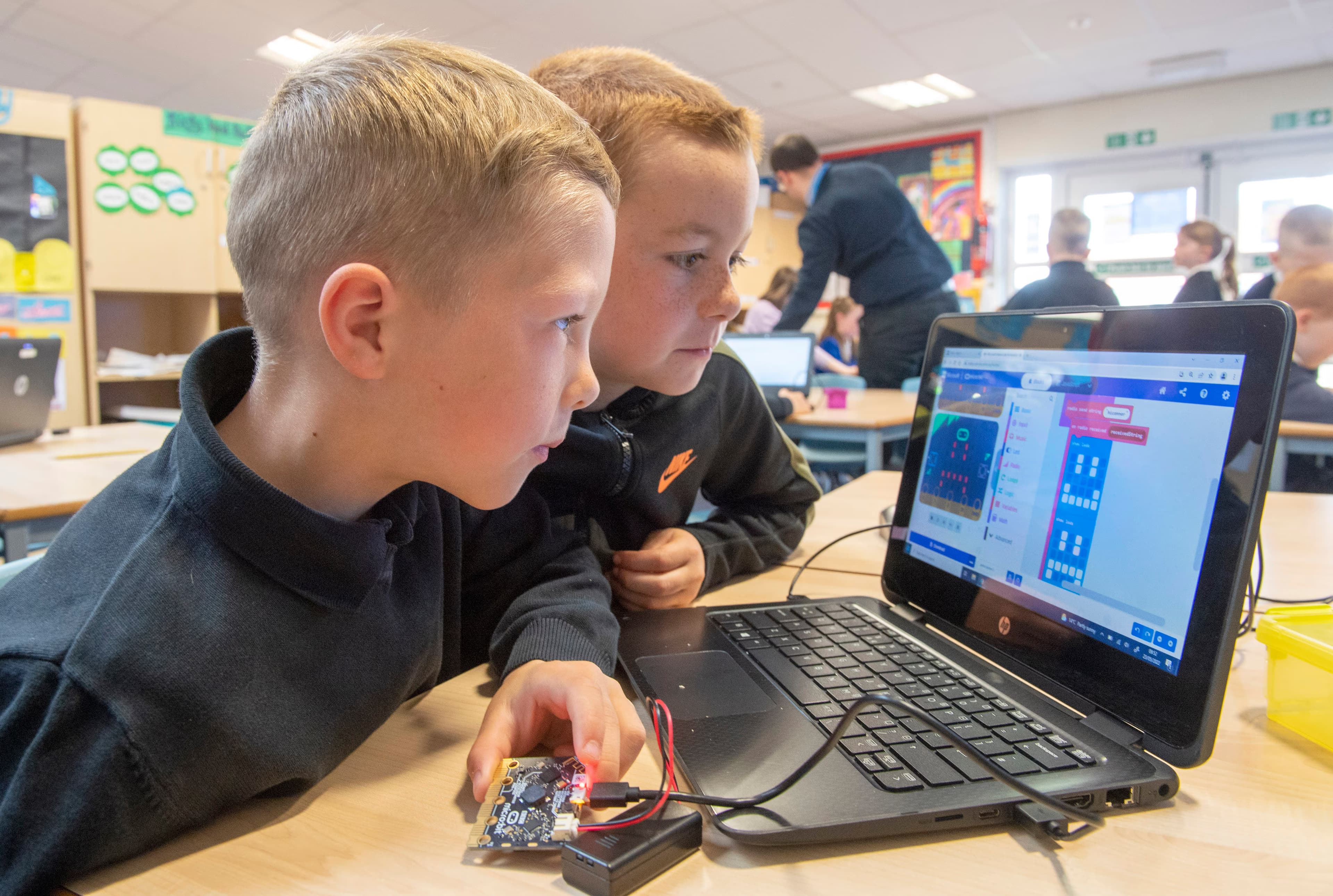 Two boys in a classroom looking at MakeCode on a laptop, one holding a micro:bit